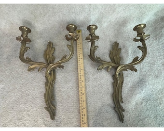 May include: Two antique brass wall sconces. Each sconce features two candle holders and a decorative, ornate leaf design. The sconces are approximately 16 inches tall and are displayed against a light-colored background.
