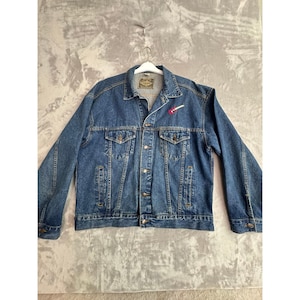 May include: A medium-wash denim jacket with a classic design. Features a button-up front, chest pockets, and side pockets. A small red and white guitar pin is attached to the lapel. The jacket is hanging on a white hanger.