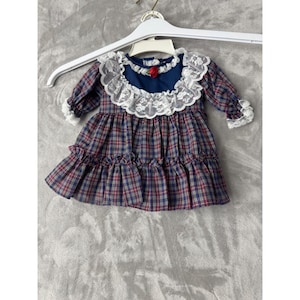 May include: A long-sleeved dress featuring a blue, red, and yellow plaid pattern. The bodice has a solid blue yoke accented with a white lace collar and a small red rose detail. The skirt and sleeves are finished with ruffled edges.