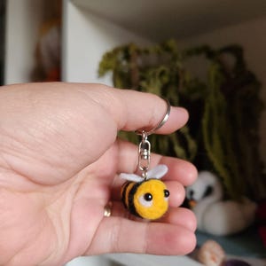 Op de afbeelding: Een gele en zwarte vilten bijensleutelhanger met witte vleugels en zwarte ogen. De bij is bevestigd aan een zilveren sleutelhanger. De sleutelhanger wordt in een hand gehouden.