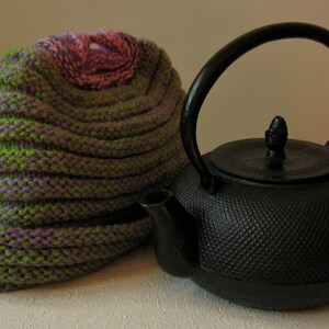 May include: A black cast iron teapot with a lid and a handle. The teapot is sitting on a white surface next to a green and purple knitted hat.