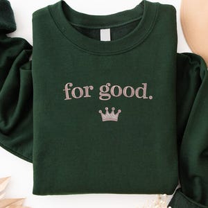 May include: A dark green sweatshirt with the phrase "for good." embroidered in a light pink color, along with a small crown. The sweatshirt has long sleeves with dark green cuffs. A tan hat is visible in the background.
