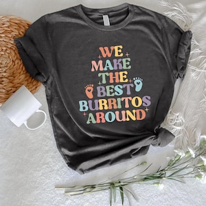 May include: Dark gray t-shirt with the text "WE MAKE THE BEST BURRITOS AROUND" in colorful, retro-style lettering. The design includes baby footprints. A white mug and woven mat are in the background.