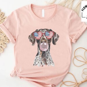 May include: A light pink short-sleeve t-shirt featuring a graphic of a brown and white spotted dog wearing heart-shaped sunglasses with an American flag pattern and blowing a pink bubblegum bubble. The shirt includes the text Flower Fashion Shirts.