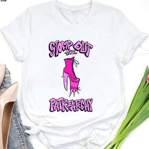 May include: White t-shirt featuring a pink high-heeled boot graphic with the text "STOMP OUT THE PATRIARCHY" in a stylized font. The shirt is displayed with a pair of light pink heels and a bouquet of pink tulips.
