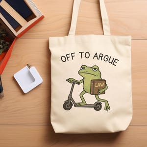 May include: A cream-colored canvas tote bag featuring a graphic of a green frog riding a scooter and holding a brown book labeled LAW. The text OFF TO ARGUE is printed in black above the frog illustration on the front of the bag.
