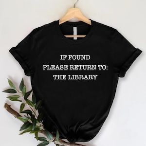 May include: Black t-shirt with the text "IF FOUND PLEASE RETURN TO: THE LIBRARY" in white. The shirt is hanging on a wooden hanger against a white background. A sprig of greenery is placed on the lower left.