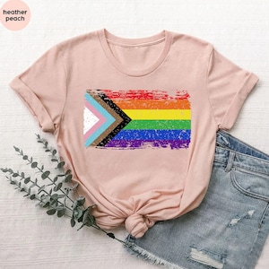 May include: A pink t-shirt with a distressed, rainbow-colored flag design. The flag includes the colors of the traditional rainbow flag, as well as black, brown, light blue, and white stripes. The flag is a symbol of inclusivity and diversity.