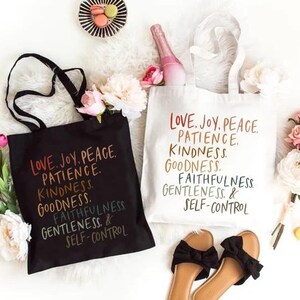 May include: Two tote bags, one black and one white, with the words "Love, Joy, Peace, Patience, Kindness, Goodness, Faithfulness, Gentleness & Self-Control" printed on them. The bags are surrounded by flowers, sandals, and a plate of macarons.