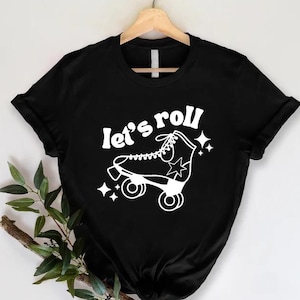 May include: Black t-shirt with a white graphic of a roller skate and the words "let's roll". The design includes stars. The shirt is hanging on a wooden hanger.