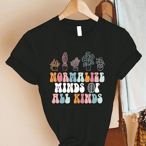 May include: Black t-shirt with a colorful graphic of potted plants and the text "NORMALIZE MINDS OF ALL KINDS" in retro font. The design includes a brain graphic. The shirt is hanging on a wooden hanger.