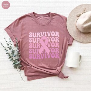 May include: Mauve-colored t-shirt with the word "SURVIVOR" in a stacked design, featuring a pink ribbon graphic. The shirt is styled with a knot at the bottom. A white mug and a beige hat are also in the image.