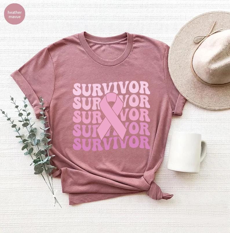 May include: Mauve-colored t-shirt with the word "SURVIVOR" in a stacked design, featuring a pink ribbon graphic. The shirt is styled with a knot at the bottom. A white mug and a beige hat are also in the image.