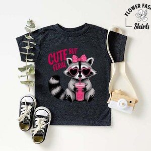 May include: Dark grey t-shirt with a cartoon raccoon wearing heart-shaped sunglasses and a pink bow. The raccoon holds a pink drink. Text on the shirt reads "Cute but Feral". Black and white trainers are at the bottom.