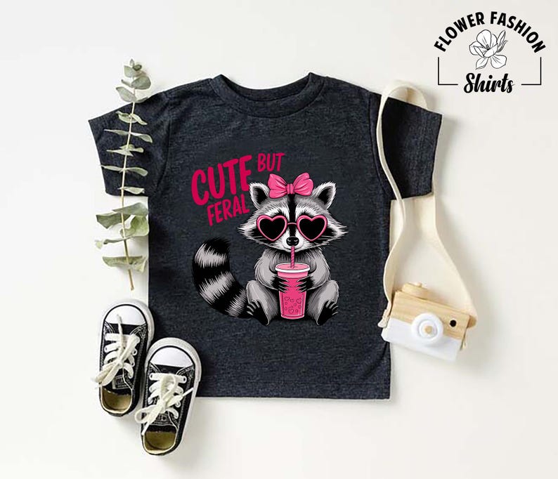 May include: Dark grey t-shirt with a cartoon raccoon wearing heart-shaped sunglasses and a pink bow. The raccoon holds a pink drink. Text on the shirt reads "Cute but Feral". Black and white trainers are at the bottom.