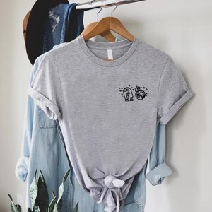 May include: A heather gray t-shirt featuring a small black graphic of two droid characters. The shirt has short sleeves and a crew neck. The shirt is hanging on a wooden hanger.