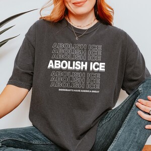 Puede incluir: Camiseta gris oscuro con la frase "ABOLISH ICE" en blanco apilado. La frase "IMMIGRANTS MAKE AMERICA GREAT" est&aacute; impresa debajo. La camiseta es de estilo casual.