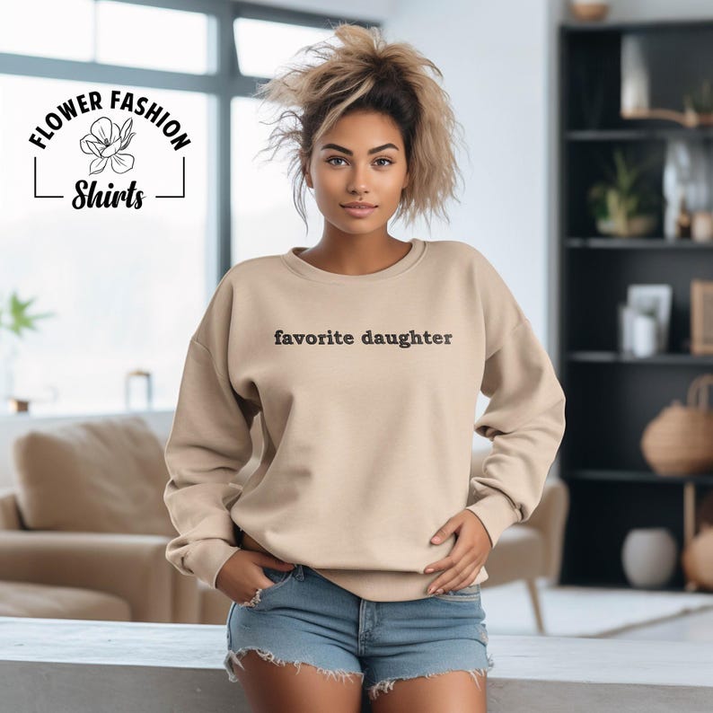 Puede incluir: Sudadera color beige con la frase "favorite daughter" en negro. La sudadera es parte de la colecci&oacute;n "Flower Fashion Shirts". La modelo lleva pantalones cortos vaqueros azules.
