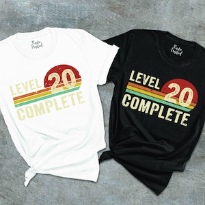 May include: Two t-shirts, one white and one black, with a retro design. The design features the text "LEVEL 20 COMPLETE" with a red circle and a rainbow-colored stripe. The shirts are likely for a birthday or milestone celebration.