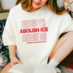 Puede incluir: Camiseta color crema con la frase "ABOLISH ICE" en rojo, repetida varias veces. La frase "IMMIGRANTS MAKE AMERICA GREAT" est&aacute; impresa debajo. La camiseta es de un material suave.