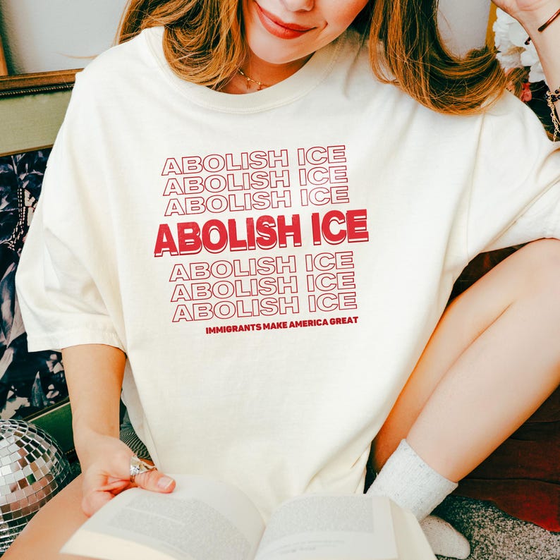 Puede incluir: Camiseta color crema con la frase "ABOLISH ICE" en rojo, repetida varias veces. La frase "IMMIGRANTS MAKE AMERICA GREAT" est&aacute; impresa debajo. La camiseta es de un material suave.