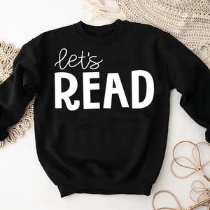 May include: Black sweatshirt with the words "let's READ" in white. The word "let's" is in a cursive font, and "READ" is in a bold, block font. The sweatshirt is a casual style, suitable for everyday wear.