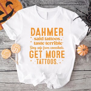 May include: White t-shirt with orange text that reads "DAHMER said tattoos taste terrible. Stay safe from cannibals. GET MORE TATTOOS." The shirt is decorated with Halloween-themed items.