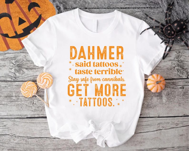 May include: White t-shirt with orange text that reads "DAHMER said tattoos taste terrible. Stay safe from cannibals. GET MORE TATTOOS." The shirt is decorated with Halloween-themed items.