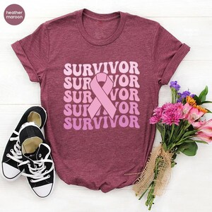 May include: Heather maroon t-shirt with the word "SURVIVOR" in a stacked, retro font, and a pink ribbon graphic. Black and white sneakers are on the left. A bouquet of pink and purple flowers is on the right.