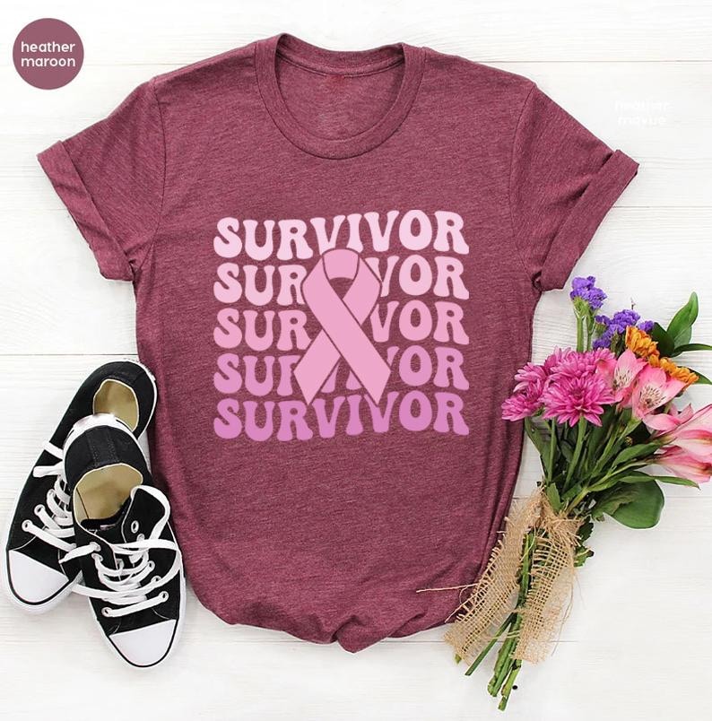 May include: Heather maroon t-shirt with the word "SURVIVOR" in a stacked, retro font, and a pink ribbon graphic. Black and white sneakers are on the left. A bouquet of pink and purple flowers is on the right.