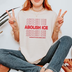 Puede incluir: Camiseta color crema con el texto "ABOLISH ICE" en rojo, apilado varias veces. La frase "IMMIGRANTS MAKE AMERICA GREAT" est&aacute; impresa debajo. La persona lleva vaqueros azules y hace el signo de la paz con ambas manos.