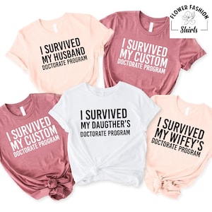 May include: A collection of five t-shirts in various colors, including white, pink, and mauve. Each shirt features the phrase "I SURVIVED" followed by a family member's relationship and "DOCTORATE PROGRAM" in bold, black text. The shirts are from Flower Fashion Shirts.