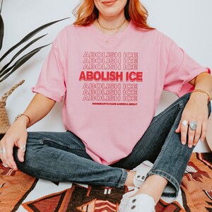 Puede incluir: Camiseta rosa con el texto "ABOLISH ICE" repetido en un dise&ntilde;o apilado. La frase "IMMIGRANTS MAKE AMERICA GREAT" est&aacute; impresa debajo. La camiseta es de color rosa liso.