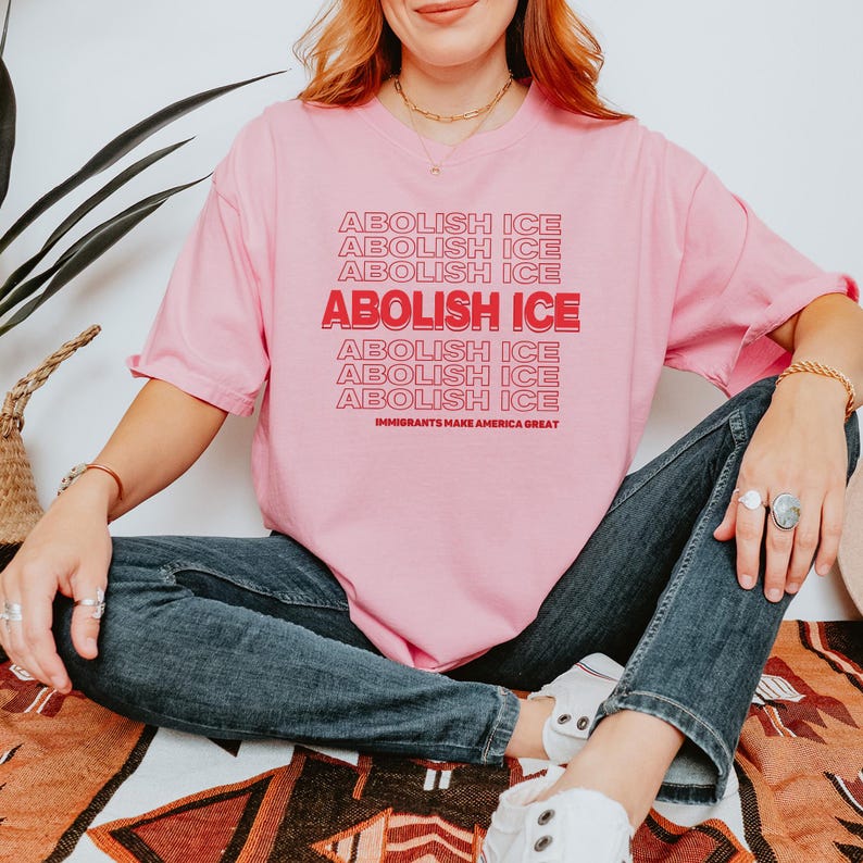 Puede incluir: Camiseta rosa con el texto "ABOLISH ICE" repetido en un dise&ntilde;o apilado. La frase "IMMIGRANTS MAKE AMERICA GREAT" est&aacute; impresa debajo. La camiseta es de color rosa liso.