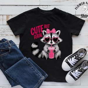 May include: Black t-shirt featuring a raccoon graphic with heart-shaped sunglasses and a pink bow, holding a pink drink. The text reads "CUTE BUT FERAL". Also includes blue jeans and black canvas trainers.