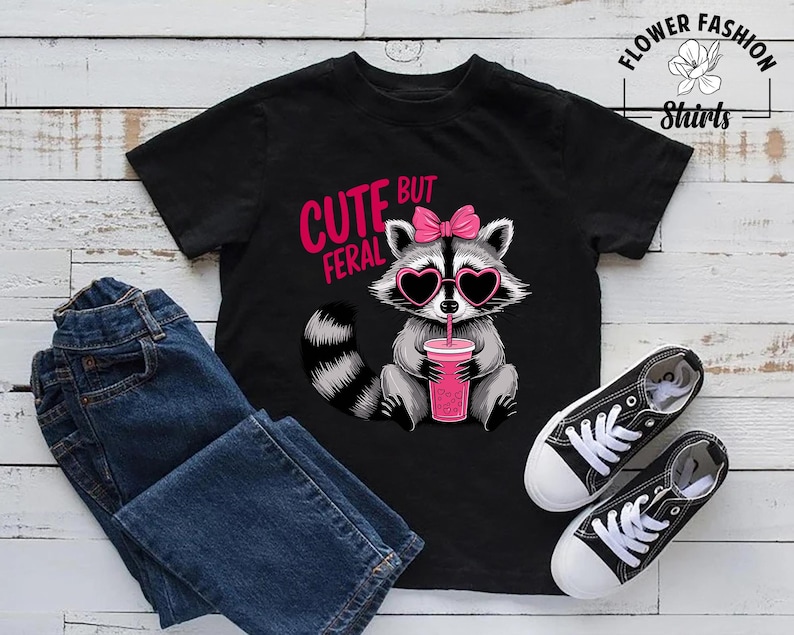 May include: Black t-shirt featuring a raccoon graphic with heart-shaped sunglasses and a pink bow, holding a pink drink. The text reads "CUTE BUT FERAL". Also includes blue jeans and black canvas trainers.