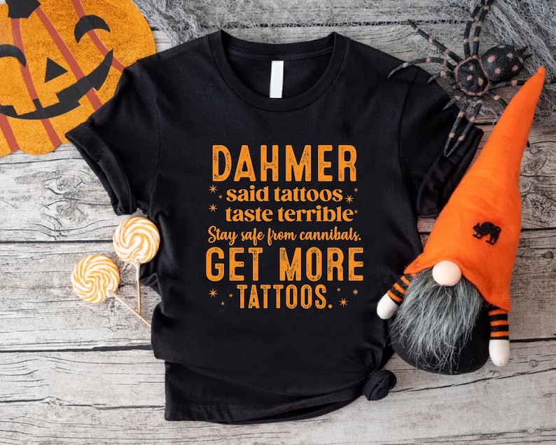 May include: Black t-shirt with orange text that reads "DAHMER said tattoos taste terrible. Stay safe from cannibals. GET MORE TATTOOS." The shirt is styled with Halloween decorations, including a pumpkin, spider, and gnome.