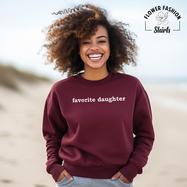 Puede incluir: Una sudadera burdeos con la frase "favorite daughter" en blanco. La sudadera es de cuello redondo. La imagen tambi&eacute;n incluye el logo "Flower Fashion Shirts".