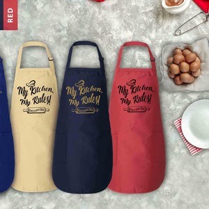 Puede incluir: Cuatro delantales en azul real, natural, marino y rojo, cada uno con el texto "My Kitchen, My Rules!". Los delantales tienen un diseño de gorro de chef y rodillo. Delantales con temática de cocina.