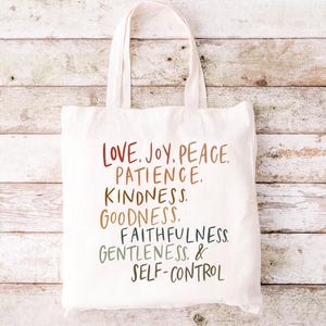 May include: A white canvas tote bag with the words "LOVE. JOY. PEACE. PATIENCE. KINDNESS. GOODNESS. FAITHFULNESS. GENTLENESS. & SELF-CONTROL" printed in a colorful, handwritten style. The bag has two handles and is set against a light wood background.