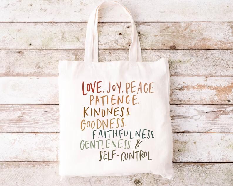 May include: A white canvas tote bag with the words "LOVE. JOY. PEACE. PATIENCE. KINDNESS. GOODNESS. FAITHFULNESS. GENTLENESS. & SELF-CONTROL" printed in a colorful, handwritten style. The bag has two handles and is set against a light wood background.