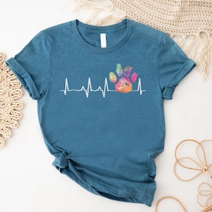 May include: A teal blue t-shirt with a white graphic of a rainbow paw print and a heartbeat line. The graphic says "Pawsitive Vibes".
