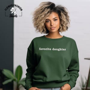 Puede incluir: Una sudadera verde oscuro con el texto blanco "favorite daughter". La sudadera es de cuello redondo. La imagen tambi&eacute;n incluye un logotipo de Flower Fashion Shirts.