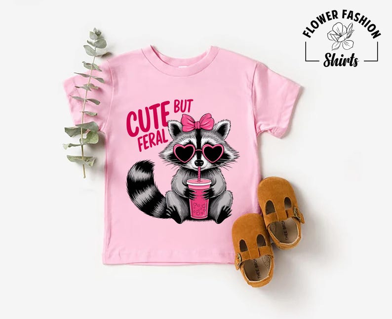 May include: A pink t-shirt featuring a raccoon wearing heart-shaped sunglasses and a pink bow, holding a pink drink. The text on the shirt reads "CUTE BUT FERAL". Brown leather baby shoes are placed next to the shirt.