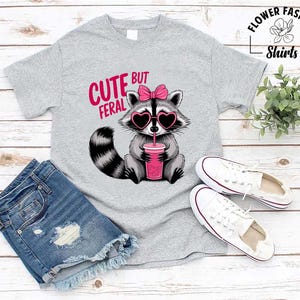 May include: A light grey t-shirt with a raccoon wearing heart-shaped sunglasses and a pink bow. The raccoon is holding a pink drink with a straw. The text "CUTE BUT FERAL" is above the raccoon. White trainers and denim shorts are also in the image.