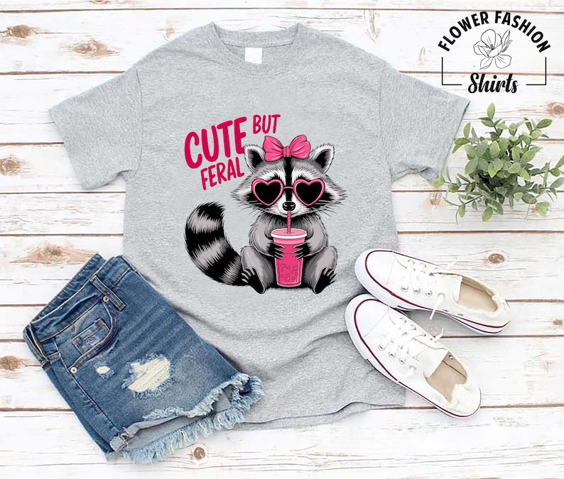 May include: A light grey t-shirt with a raccoon wearing heart-shaped sunglasses and a pink bow. The raccoon is holding a pink drink with a straw. The text "CUTE BUT FERAL" is above the raccoon. White trainers and denim shorts are also in the image.