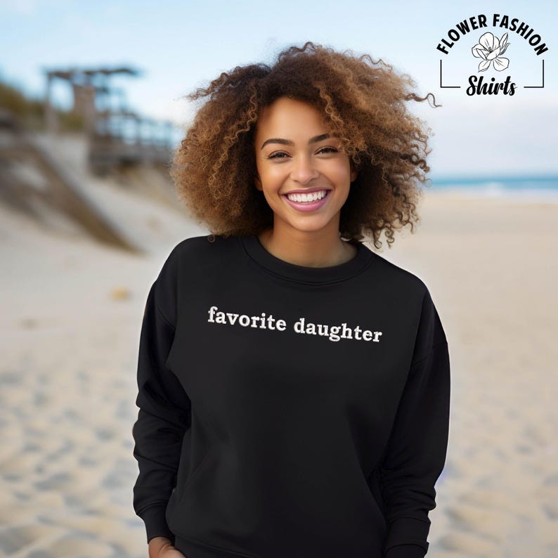 Puede incluir: Sudadera negra con la frase "favorite daughter" en blanco. La sudadera es de cuello redondo. La imagen tambi&eacute;n incluye el logotipo de Flower Fashion Shirts en la esquina superior derecha. El fondo es una escena de playa con arena y agua.