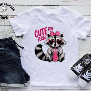May include: White t-shirt featuring a raccoon graphic with heart-shaped sunglasses and a pink bow, holding a pink drink. The text "CUTE BUT FERAL" is above the raccoon. The shirt is styled with denim shorts and black trainers.
