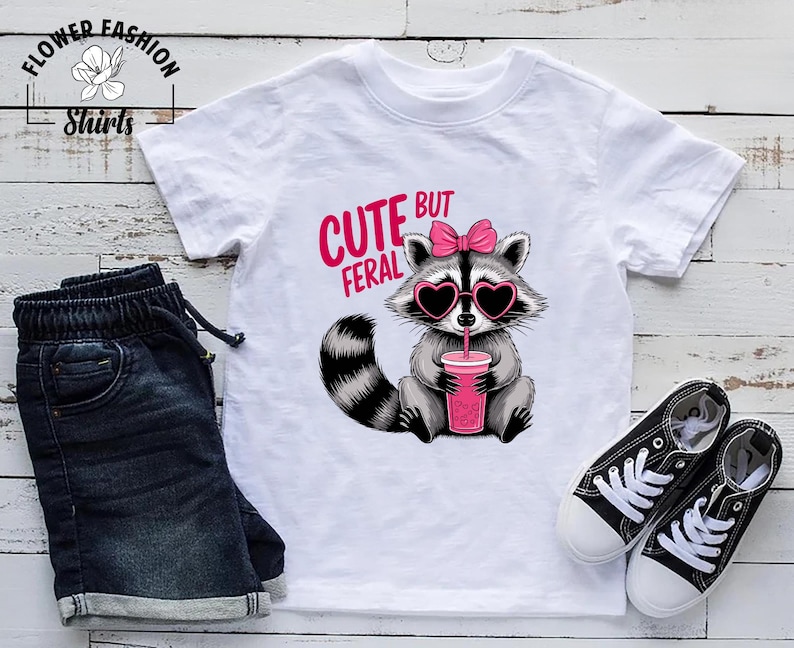 May include: White t-shirt featuring a raccoon graphic with heart-shaped sunglasses and a pink bow, holding a pink drink. The text "CUTE BUT FERAL" is above the raccoon. The shirt is styled with denim shorts and black trainers.