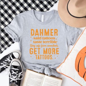 May include: A gray t-shirt with orange text that reads "DAHMER said tattoos taste terrible. Stay safe from cannibals. GET MORE TATTOOS." The shirt is paired with black sneakers, a tan hat, and a pumpkin pillow.